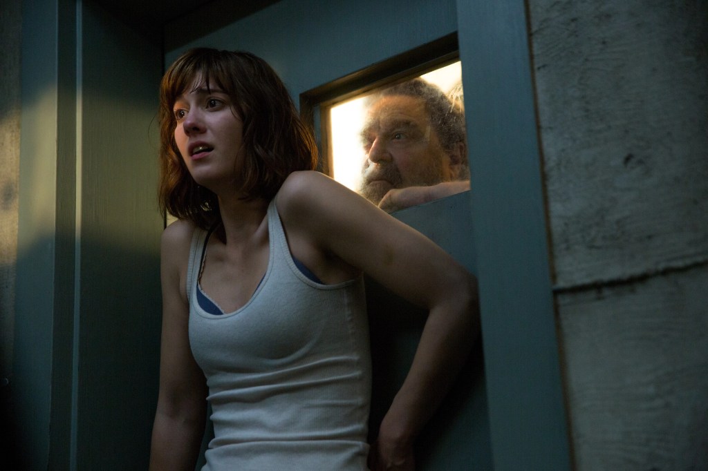 A scared woman stands in front of a door as a threatening man leers at her from a transom window.